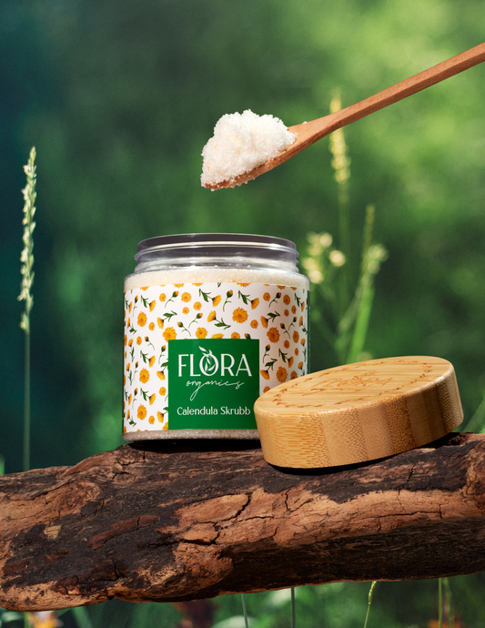 Calendula body scrub from Flora Organics in open jar on tree bark, with wooden lid and spoonful of scrub held above