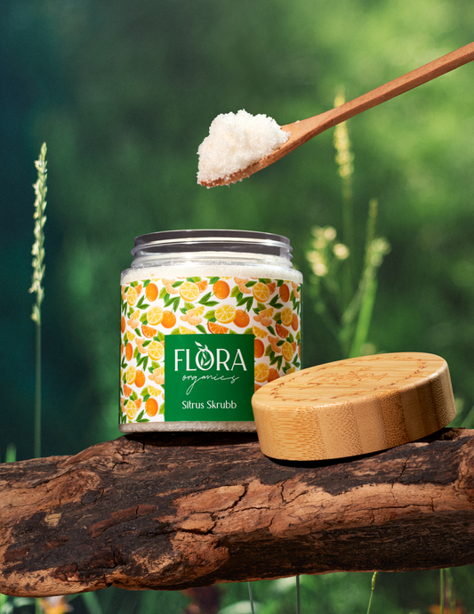 Natural and Organic Citrus body scrub from Flora Organics in open jar on tree bark, with wooden lid and spoonful of scrub held above