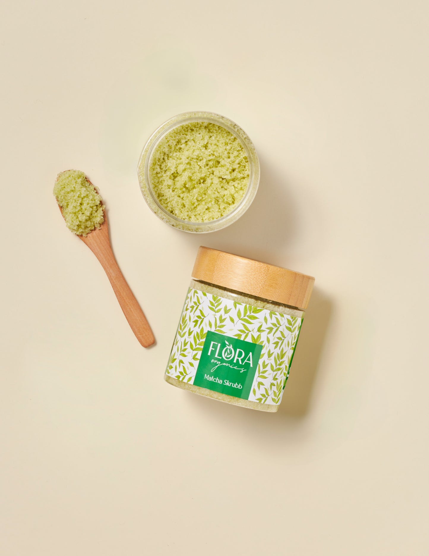 Top view of Flora Organics Matcha Scrub in glass jar with bamboo lid, next to wooden spoon and open bowl of green scrub on beige background