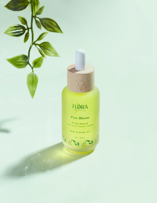 Flora Organics Pure Bloom Oil bottle with dropper cap on glossy green surface, with hanging plant leaves above