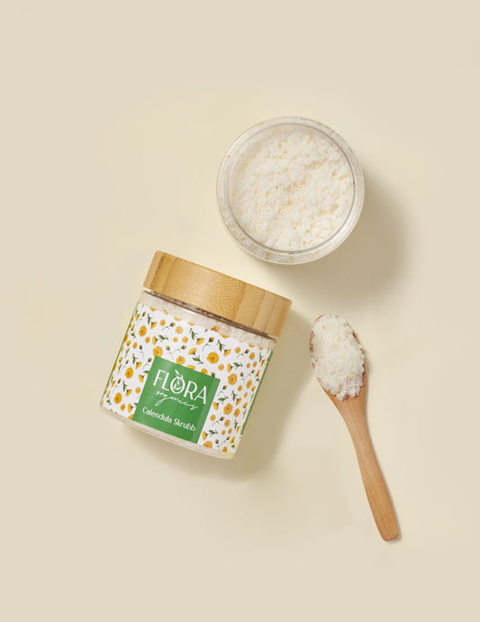 Top view of Flora Organics Calendula Scrub in glass jar with bamboo lid, wooden spoon and bowl of soft exfoliant on beige background