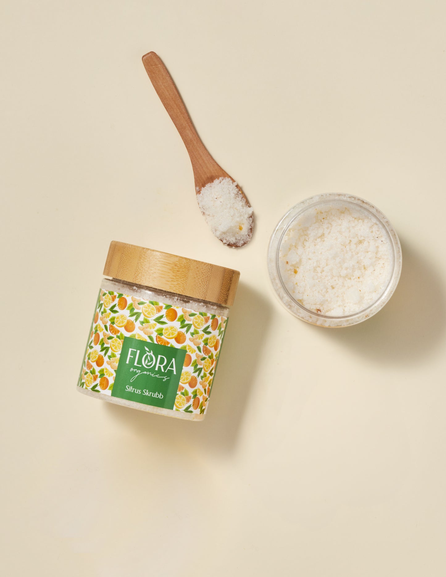 Top view of Flora Organics Citrus Scrub with bamboo lid, wooden spoon, and glass bowl of exfoliating salts on beige background
