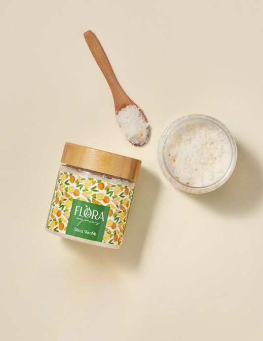 Top view of Flora Organics Citrus Scrub with bamboo lid, wooden spoon, and glass bowl of exfoliating salts on beige background
