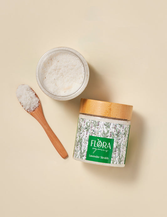 Top view of Flora Organics Lavender Scrub in glass jar with bamboo lid, next to wooden spoon and open bowl of exfoliant on beige background