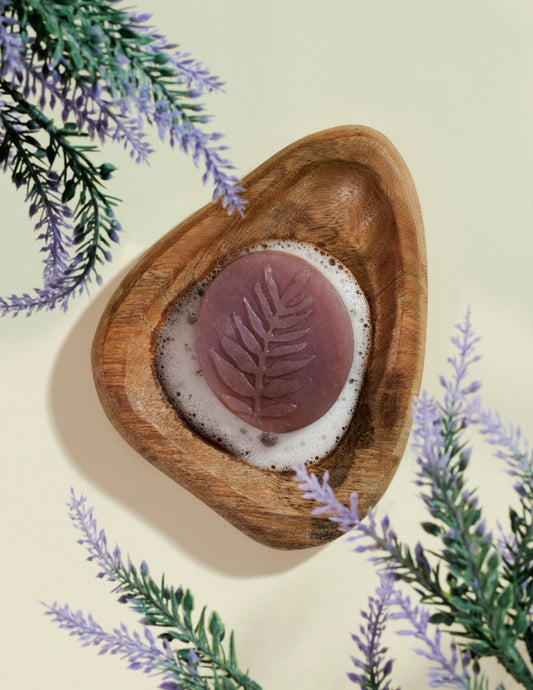Lavender soap bar with fern imprint foaming in wooden dish, surrounded by lavender sprigs on cream background