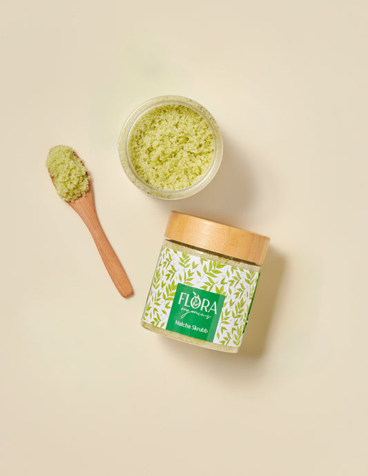 Top view of Flora Organics Matcha Scrub in glass jar with bamboo lid, next to wooden spoon and open bowl of green scrub on beige background
